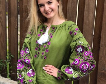 Puede incluir: Una blusa de lino verde con diseños florales bordados en morado y negro. La blusa tiene un borla blanca en el escote y mangas largas.