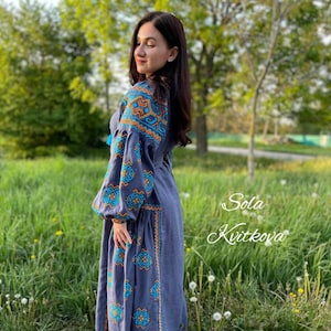 May include: A woman wearing a long, gray linen dress with blue and orange embroidered patterns. The dress has a loose fit and long sleeves. The woman is standing in a grassy field with trees in the background. The text "Sola Kvitkova" is written in cursive on the right side of the image.