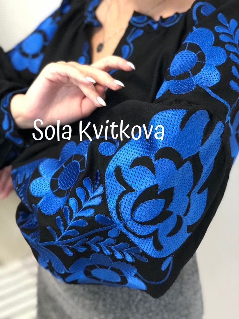 Op de afbeelding: Een zwarte geborduurde blouse met blauwe bloemenmotieven. De tekst "Sola Kvitkova" is zichtbaar op de voorkant van de blouse.