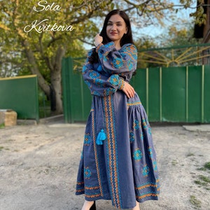 May include: A woman wearing a long, blue embroidered dress with orange and blue accents. The dress has a blue tassel hanging from the waist. The woman is standing in front of a green gate and a tree. The text "Sola Kvitkova" is written in white on a black background in the top left corner of the image.
