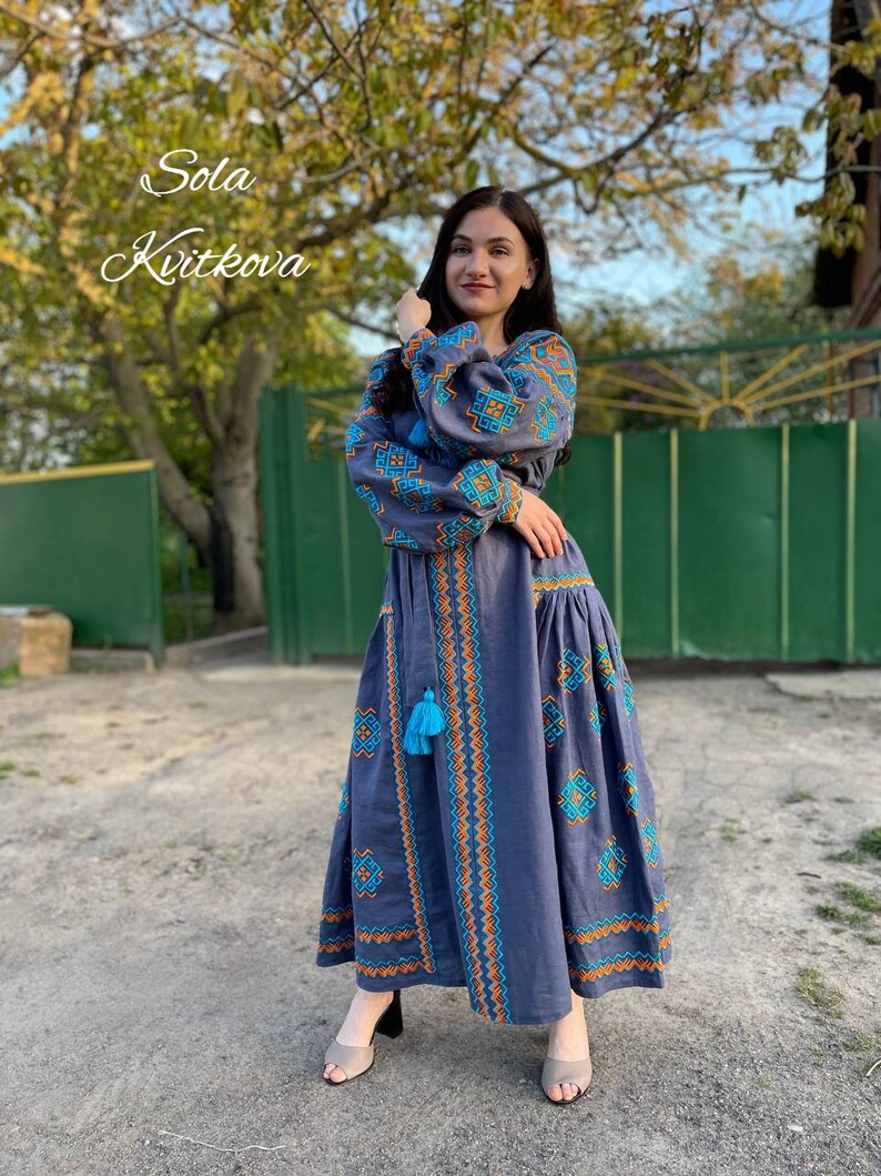 May include: A woman wearing a long, blue embroidered dress with orange and blue accents. The dress has a blue tassel hanging from the waist. The woman is standing in front of a green gate and a tree. The text "Sola Kvitkova" is written in white on a black background in the top left corner of the image.