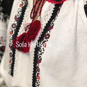 May include: White linen blouse with black and red embroidered details. The blouse has a red tassel hanging from the neckline. The text "Sola Kvitkova" is embroidered on the front of the blouse.
