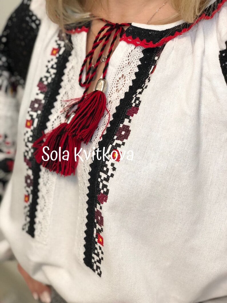 May include: White linen blouse with black and red embroidered details. The blouse has a red tassel hanging from the neckline. The text "Sola Kvitkova" is embroidered on the front of the blouse.