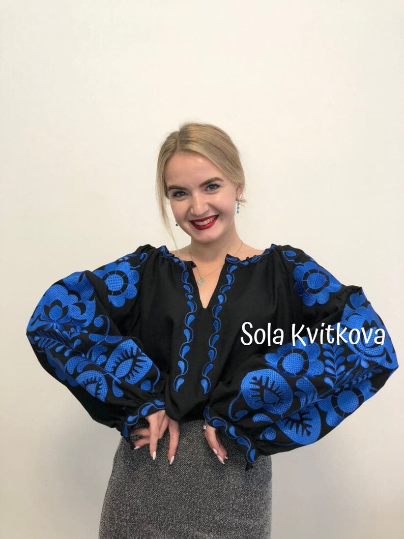Op de afbeelding: Een zwarte geborduurde blouse met blauwe bloemenpatronen. De blouse heeft een V-hals en lange mouwen. De tekst "Sola Kvitkova" is zichtbaar in de rechterbenedenhoek van de afbeelding.