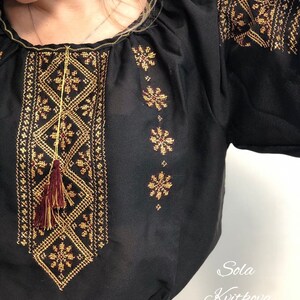 Pu&ograve; includere: Blusa nera in chiffon con ricami dorati e bordeaux. Il ricamo presenta motivi geometrici e nappe. La blusa ha un collo rotondo e maniche corte.