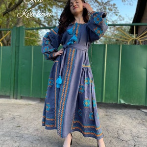 May include: A long, blue linen dress with embroidered geometric patterns in orange and turquoise. The dress has a cinched waist with a matching belt and features tassels on the sleeves and hem.