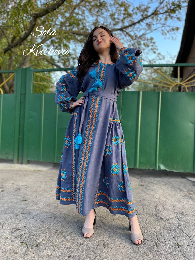 May include: A long, blue linen dress with embroidered geometric patterns in orange and turquoise. The dress has a cinched waist with a matching belt and features tassels on the sleeves and hem.
