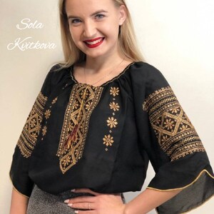 Pu&ograve; includere: Una blusa nera ricamata con filo dorato e un fiocco rosso. La blusa ha una scollatura rotonda e maniche a 3/4. Il testo "Sola Kvitkova" &egrave; visibile nell'angolo in alto a sinistra dell'immagine.