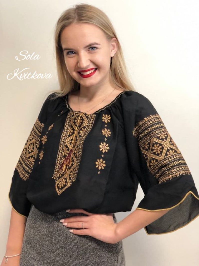 Pu&ograve; includere: Una blusa nera ricamata con filo dorato e un fiocco rosso. La blusa ha una scollatura rotonda e maniche a 3/4. Il testo "Sola Kvitkova" &egrave; visibile nell'angolo in alto a sinistra dell'immagine.