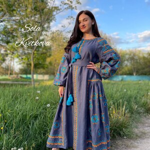 May include: A woman wearing a long, gray linen dress with blue and orange embroidery. The dress has a belt and long sleeves with tassels. The dress is called "Sola Kvitkova".