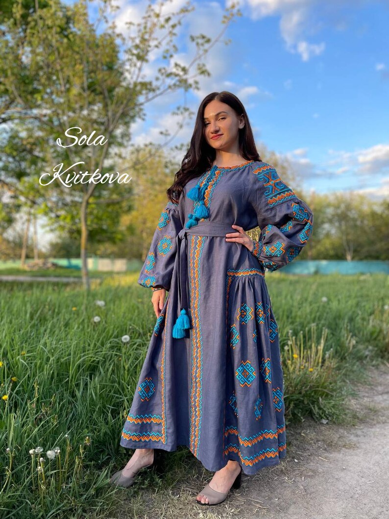 May include: A woman wearing a long, gray linen dress with blue and orange embroidery. The dress has a belt and long sleeves with tassels. The dress is called "Sola Kvitkova".