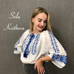 May include: A white blouse with blue embroidery and a tassel. The blouse has a round neckline and long sleeves. The embroidery is a traditional Ukrainian design.