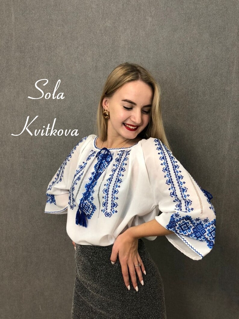 May include: A white blouse with blue embroidery and a tassel. The blouse has a round neckline and long sleeves. The embroidery is a traditional Ukrainian design.