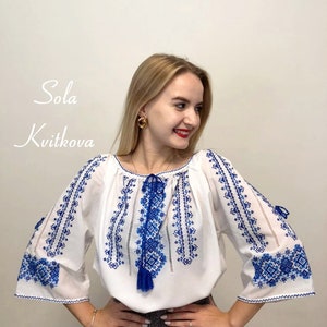 May include: A white blouse with blue embroidery. The embroidery features a geometric pattern. The blouse has a gathered neckline and long sleeves.