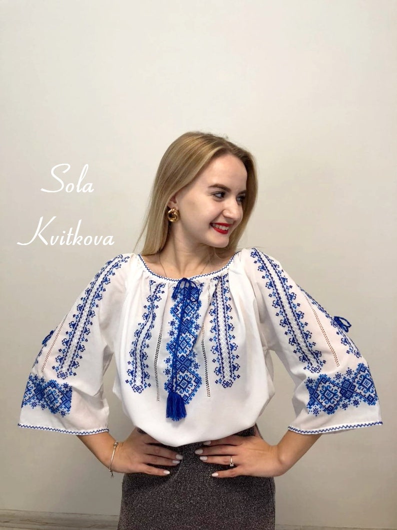 May include: A white blouse with blue embroidery. The embroidery features a geometric pattern. The blouse has a gathered neckline and long sleeves.