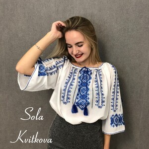 May include: A white blouse with blue embroidered floral patterns and tassels. The blouse has a loose fit and a round neckline. The text "Sola Kvitkova" is visible in the image.