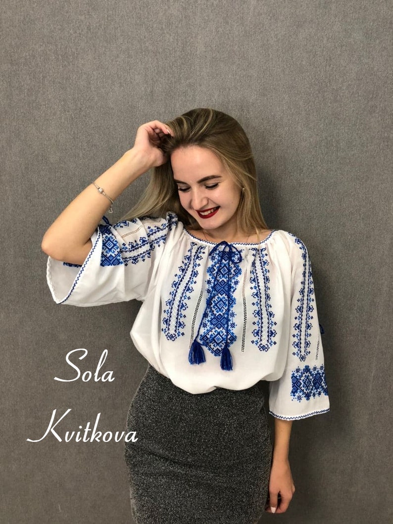 May include: A white blouse with blue embroidered floral patterns and tassels. The blouse has a loose fit and a round neckline. The text "Sola Kvitkova" is visible in the image.