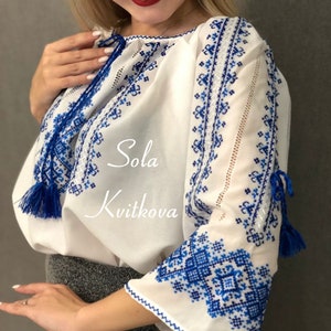 May include: White embroidered blouse with blue floral patterns and tassels. The blouse has a round neckline and long sleeves. The text "Sola Kvitkova" is embroidered on the front of the blouse.