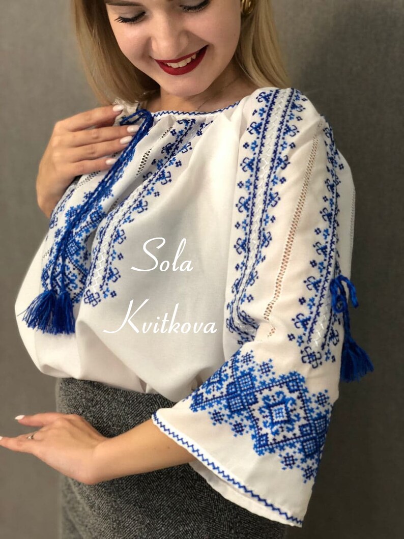 May include: White embroidered blouse with blue floral patterns and tassels. The blouse has a round neckline and long sleeves. The text "Sola Kvitkova" is embroidered on the front of the blouse.