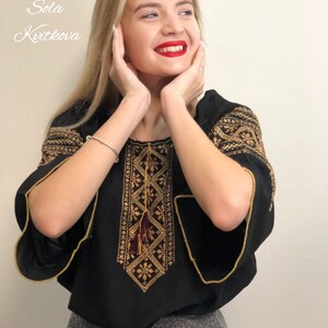 Pu&ograve; includere: Una blusa nera ricamata con filo dorato e un fiocco. La blusa ha una scollatura rotonda e maniche corte. La modella indossa una gonna grigia.