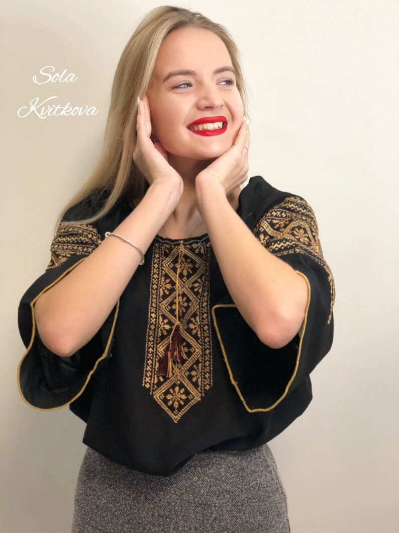 Pu&ograve; includere: Una blusa nera ricamata con filo dorato e un fiocco. La blusa ha una scollatura rotonda e maniche corte. La modella indossa una gonna grigia.