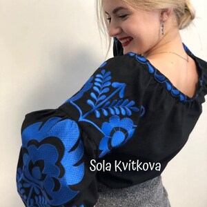 Op de afbeelding: Een zwart blouse met blauwe geborduurde bloemenmotieven. De blouse heeft een ronde halslijn en lange, pofmouwen. De tekst "Sola Kvitkova" is zichtbaar op de blouse.