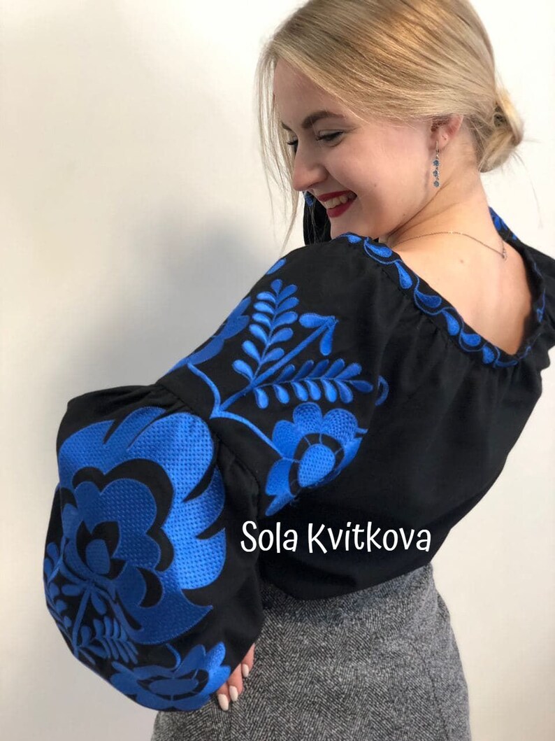 Op de afbeelding: Een zwart blouse met blauwe geborduurde bloemenmotieven. De blouse heeft een ronde halslijn en lange, pofmouwen. De tekst "Sola Kvitkova" is zichtbaar op de blouse.