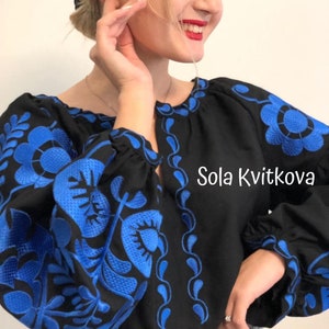 Op de afbeelding: Een zwart blouse met blauwe geborduurde bloemenpatronen. De blouse heeft een ronde hals en lange mouwen. De tekst "Sola Kvitkova" is geborduurd aan de rechterkant van de blouse.