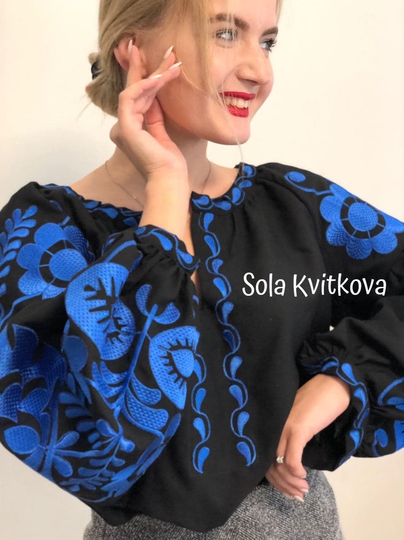 Op de afbeelding: Een zwart blouse met blauwe geborduurde bloemenpatronen. De blouse heeft een ronde hals en lange mouwen. De tekst "Sola Kvitkova" is geborduurd aan de rechterkant van de blouse.