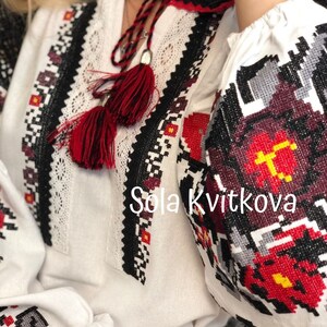 May include: White embroidered blouse with red and black geometric patterns. The blouse has a red tassel tie and the text "Sola Kvitkova" is embroidered on the front.