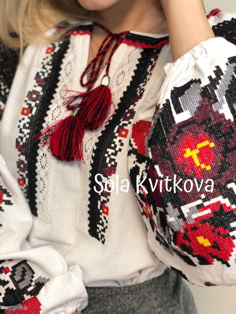 May include: White embroidered blouse with red and black geometric patterns. The blouse has a red tassel tie and the text "Sola Kvitkova" is embroidered on the front.