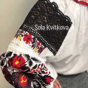 May include: White embroidered blouse with black lace detailing on the sleeves. The embroidery features red, black, and yellow floral patterns. The text "Sola Kvitkova" is visible on the blouse.