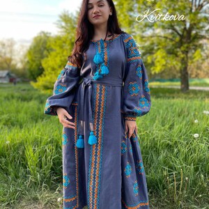 May include: A woman wearing a long, blue linen dress with embroidered details in orange and turquoise. The dress has a belt and long sleeves. The woman is standing in a grassy field with trees in the background. The text "Sola Kvitkova" is written in cursive on the right side of the image.