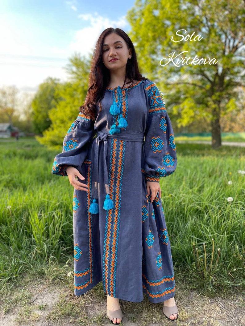 May include: A woman wearing a long, blue linen dress with embroidered details in orange and turquoise. The dress has a belt and long sleeves. The woman is standing in a grassy field with trees in the background. The text "Sola Kvitkova" is written in cursive on the right side of the image.