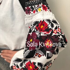 May include: White embroidered blouse with black lace sleeves and red and black floral embroidery. The embroidery features a repeating pattern of flowers and leaves. The text "Sola Kvitkova" is embroidered on the sleeve.