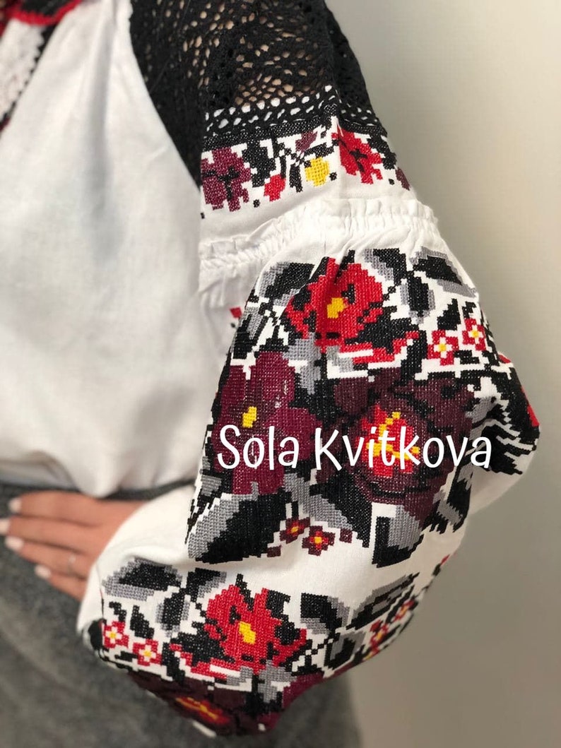 May include: White embroidered blouse with black lace sleeves and red and black floral embroidery. The embroidery features a repeating pattern of flowers and leaves. The text "Sola Kvitkova" is embroidered on the sleeve.