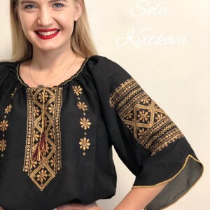 Pu&ograve; includere: Una blusa nera ricamata con filo dorato e un fiocco marrone. La blusa ha un collo a sbuffo e maniche 3/4. Il testo "Sola Kvitkova" &egrave; visibile nell'angolo in alto a destra dell'immagine.