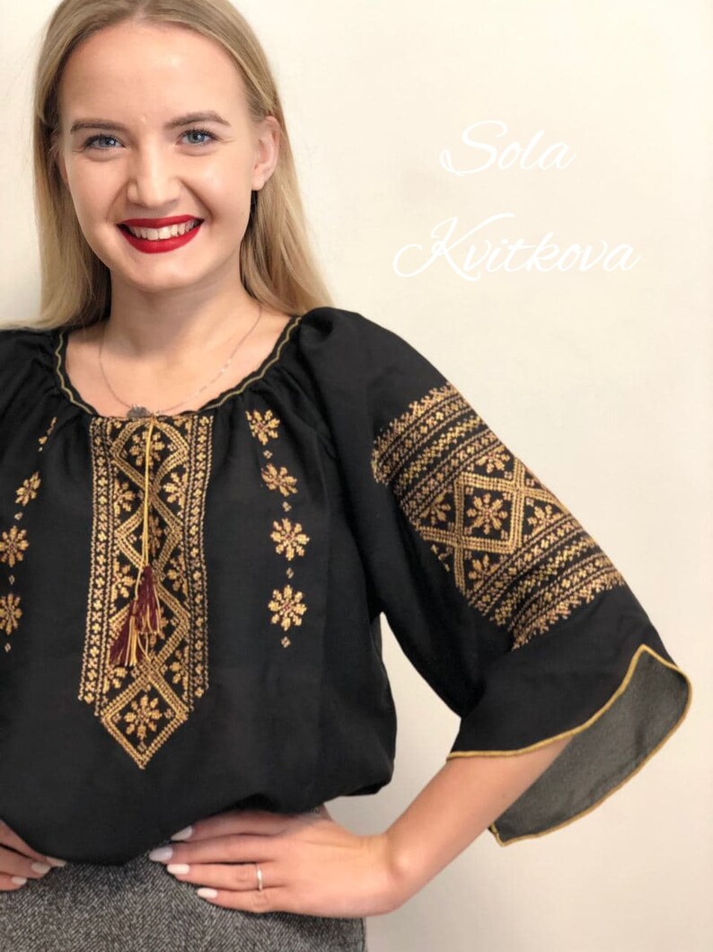 Pu&ograve; includere: Una blusa nera ricamata con filo dorato e un fiocco marrone. La blusa ha un collo a sbuffo e maniche 3/4. Il testo "Sola Kvitkova" &egrave; visibile nell'angolo in alto a destra dell'immagine.