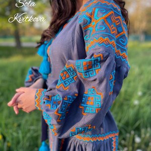 May include: A gray linen dress with blue and orange embroidery. The embroidery features geometric patterns. The dress has a gathered skirt and long sleeves.