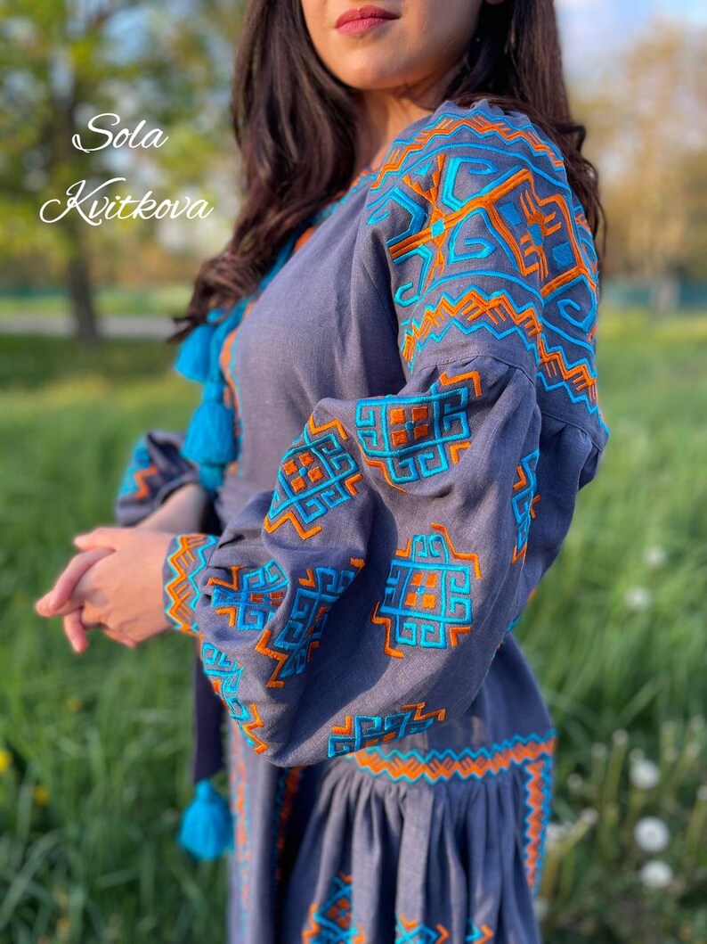May include: A gray linen dress with blue and orange embroidery. The embroidery features geometric patterns. The dress has a gathered skirt and long sleeves.