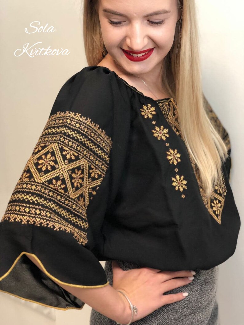 Pu&ograve; includere: Una blusa nera con ricami dorati. Il ricamo presenta un motivo geometrico e motivi floreali. La blusa ha un collo a volant e maniche lunghe.