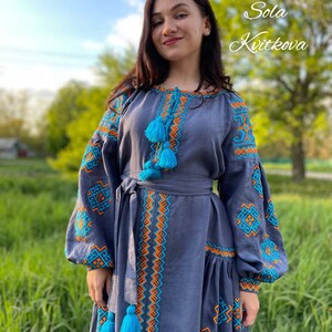 May include: A woman wearing a long, blue linen dress with embroidered details in orange and turquoise. The dress has a fitted bodice and a flowing skirt with a gathered hem. The dress features a belt and tassels.