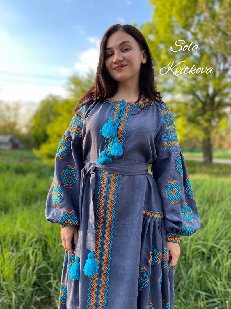 May include: A woman wearing a long, blue linen dress with embroidered details in orange and turquoise. The dress has a fitted bodice and a flowing skirt with a gathered hem. The dress features a belt and tassels.