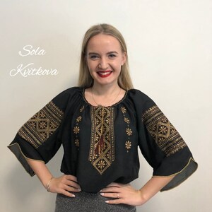 Pu&ograve; includere: Una blusa nera con ricami dorati che presentano un motivo geometrico. La blusa ha una scollatura rotonda e maniche a tre quarti con bordi dorati.