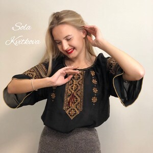 Pu&ograve; includere: Una blusa nera ricamata con filo dorato e marrone. La blusa ha un fiocco che pende dal ricamo. La modella indossa una gonna grigia.