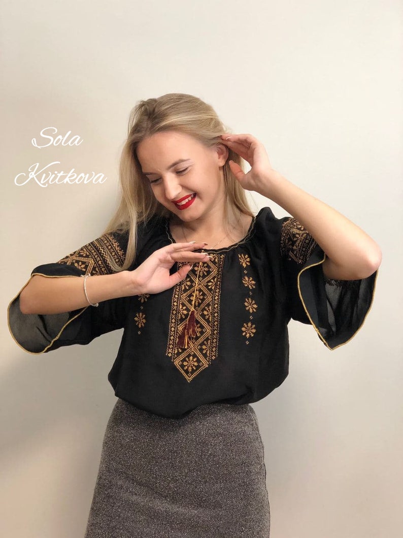 Pu&ograve; includere: Una blusa nera ricamata con filo dorato e marrone. La blusa ha un fiocco che pende dal ricamo. La modella indossa una gonna grigia.