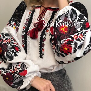 May include: White embroidered blouse with black and red floral patterns. The blouse has long sleeves and a round neckline. The text "Sola Kvitkova" is visible on the front of the blouse.