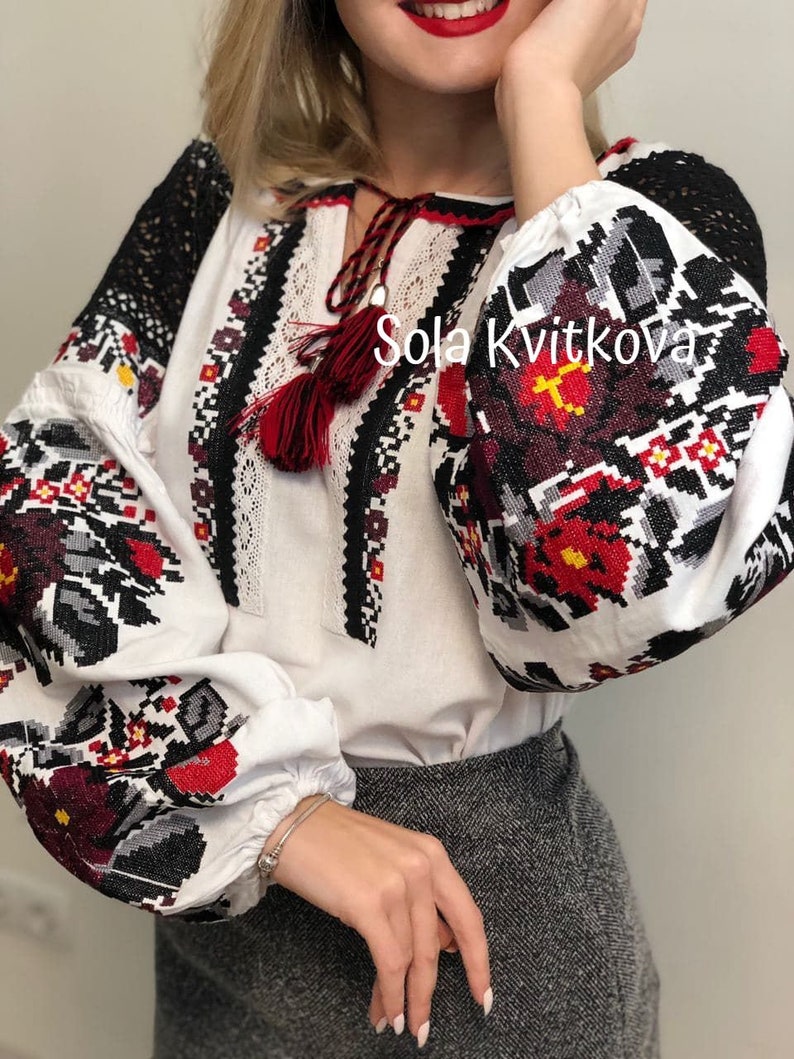 May include: White embroidered blouse with black and red floral patterns. The blouse has long sleeves and a round neckline. The text "Sola Kvitkova" is visible on the front of the blouse.