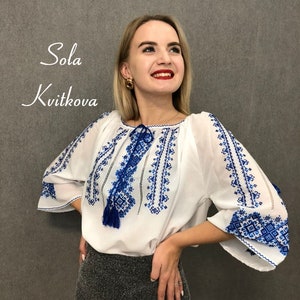May include: A white blouse with blue embroidered details and a tassel. The blouse has a round neckline and 3/4 sleeves. The model is wearing a black skirt.