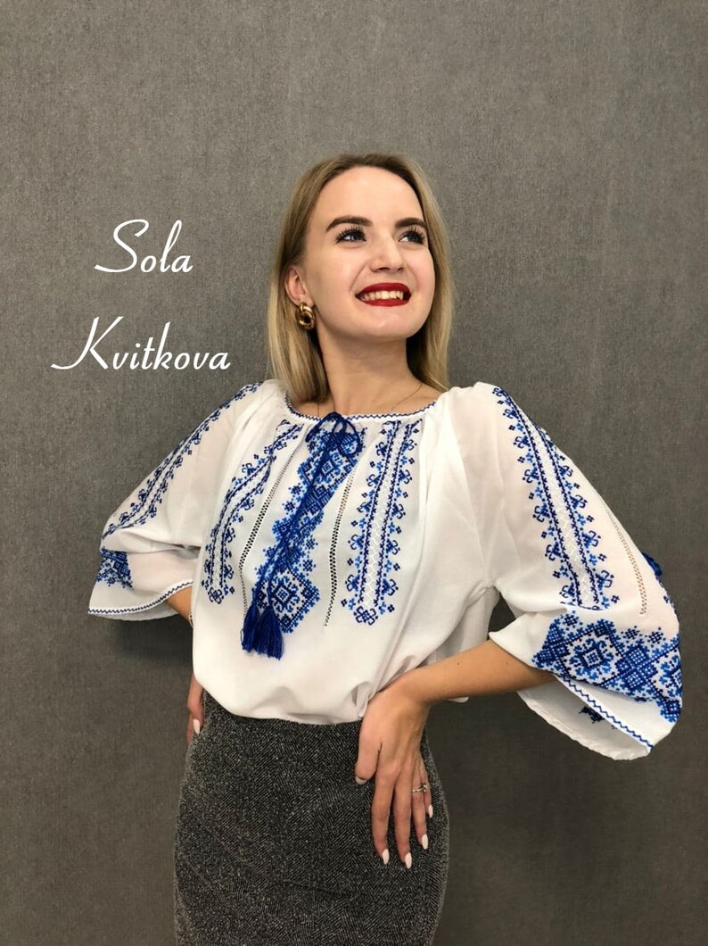 May include: A white blouse with blue embroidered details and a tassel. The blouse has a round neckline and 3/4 sleeves. The model is wearing a black skirt.